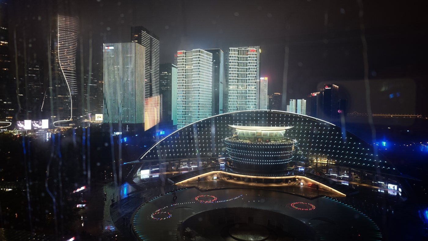 Hangzhou at night through rain-streaked glass, a dome building lit blue and gold below and lit towers behind it