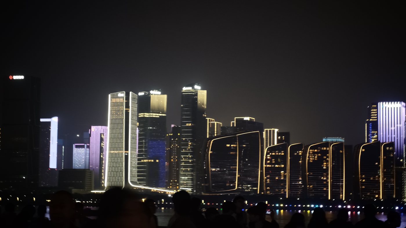 Lit towers on the far side of the water in Hangzhou at night, with glowing outlines and a crowd along the front