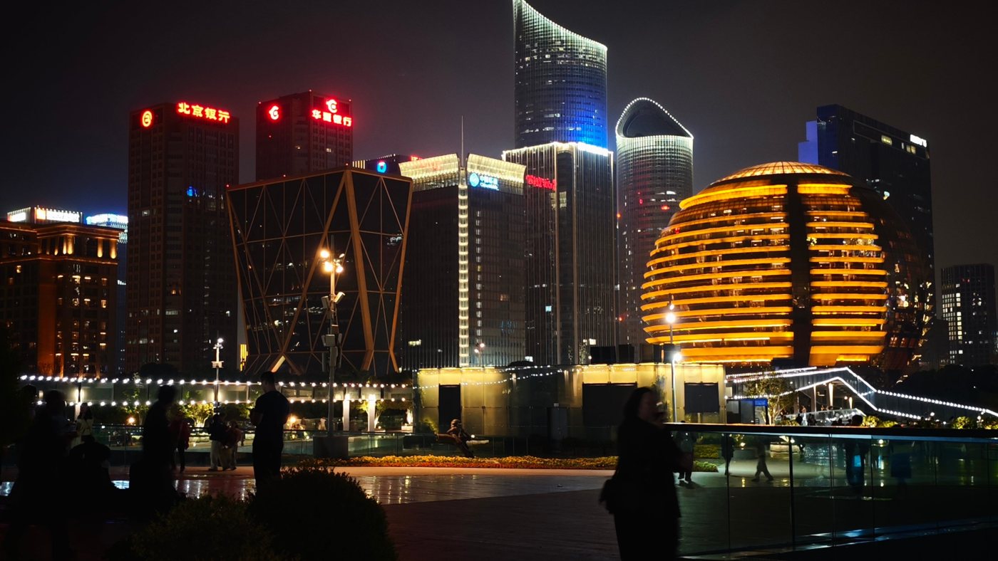 The golden globe of InterContinental Hangzhou ICC lit orange at night beside lit towers, with people on the promenade in front