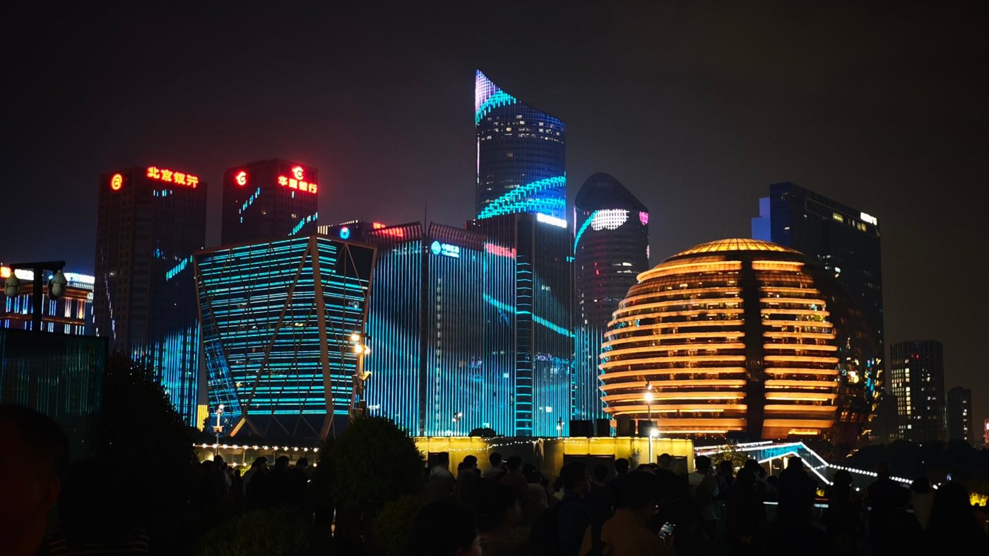 The golden globe of InterContinental Hangzhou ICC next to towers lit blue by a light show, with a crowd watching in front