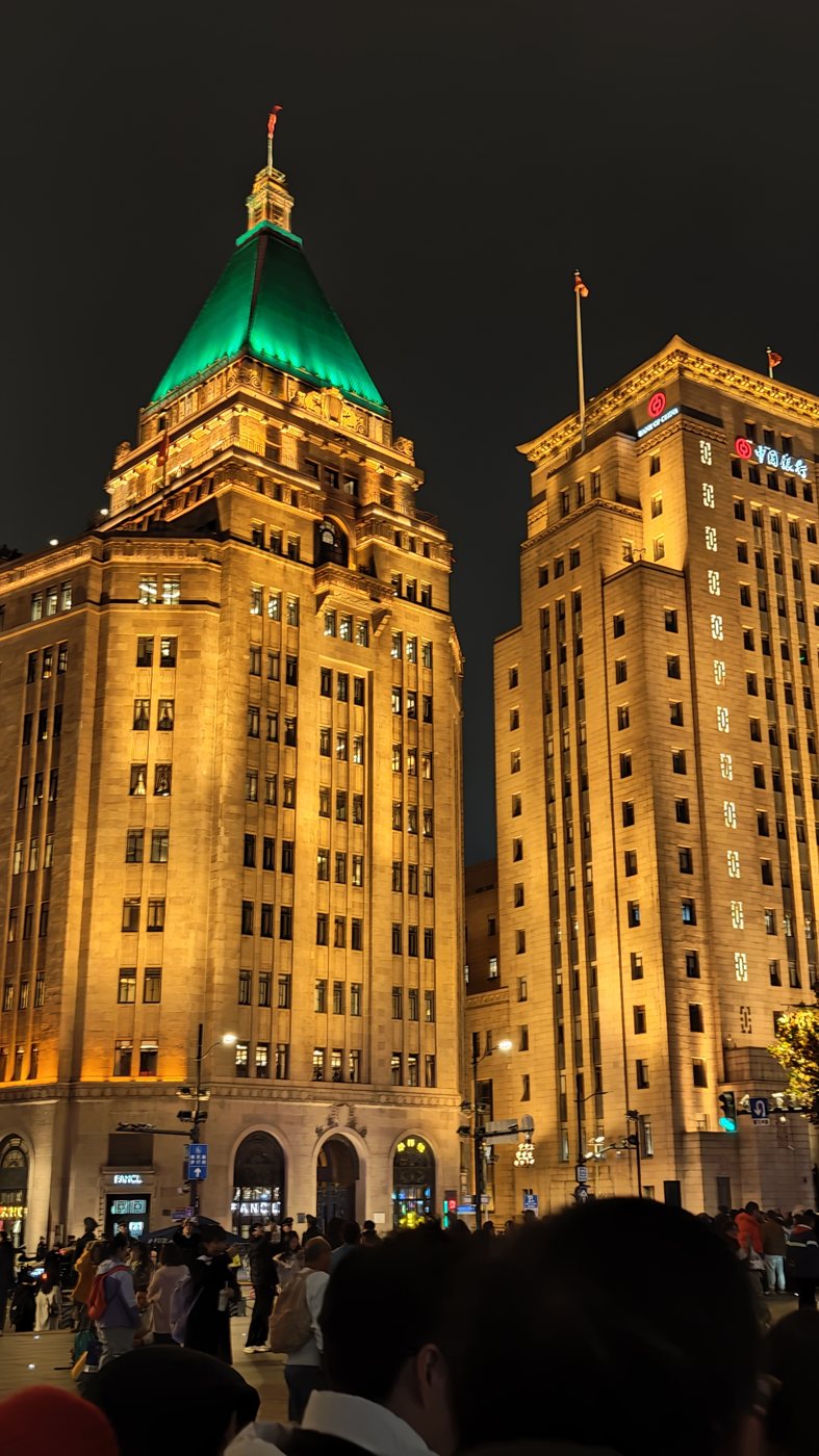 The green-roofed Peace Hotel and the tower beside it on the Bund in Shanghai, lit gold at night with a crowd below