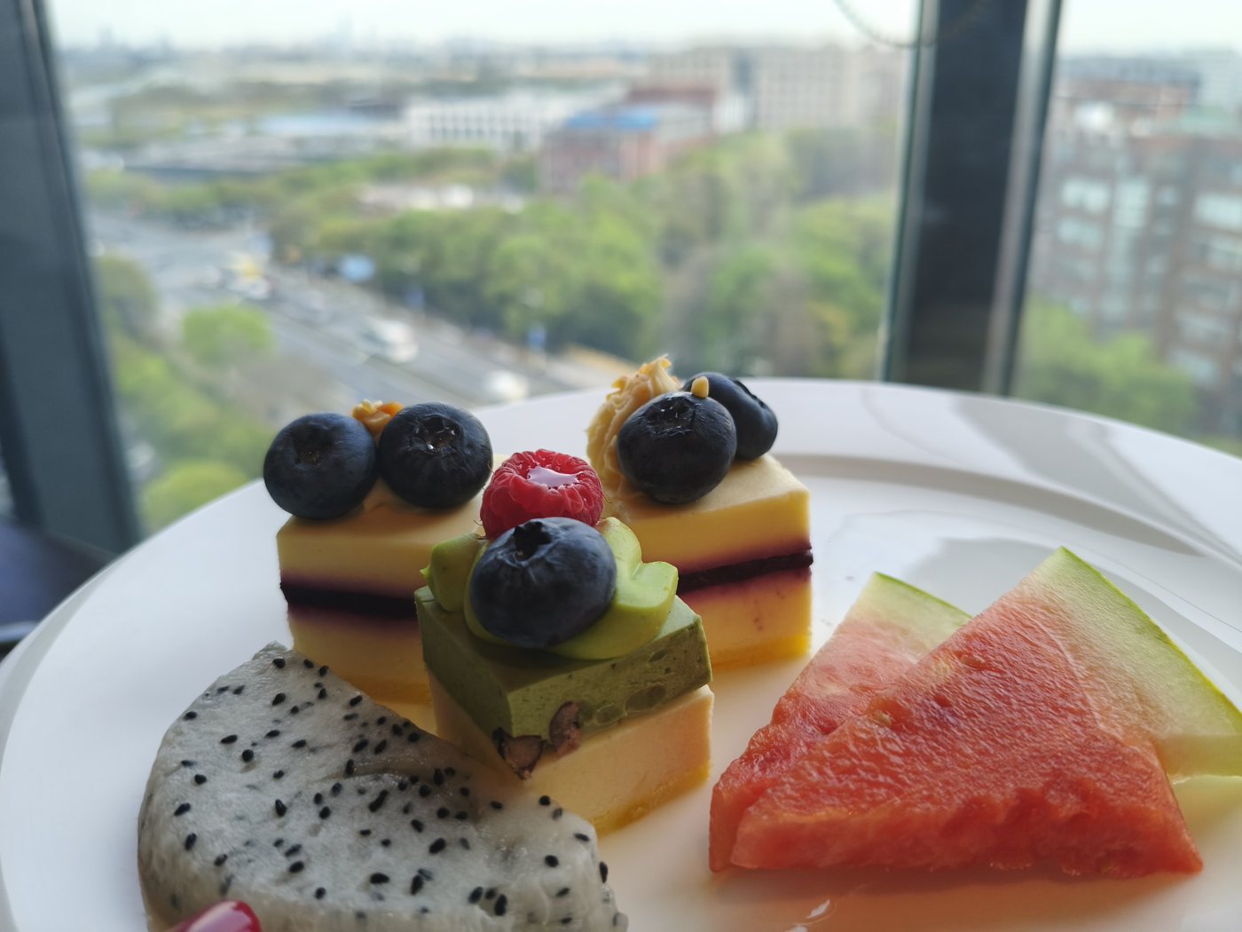 A plate of layered cake squares with blueberries, dragon fruit and watermelon in the voco Shanghai Hongqiao Hub lounge, green city view behind
