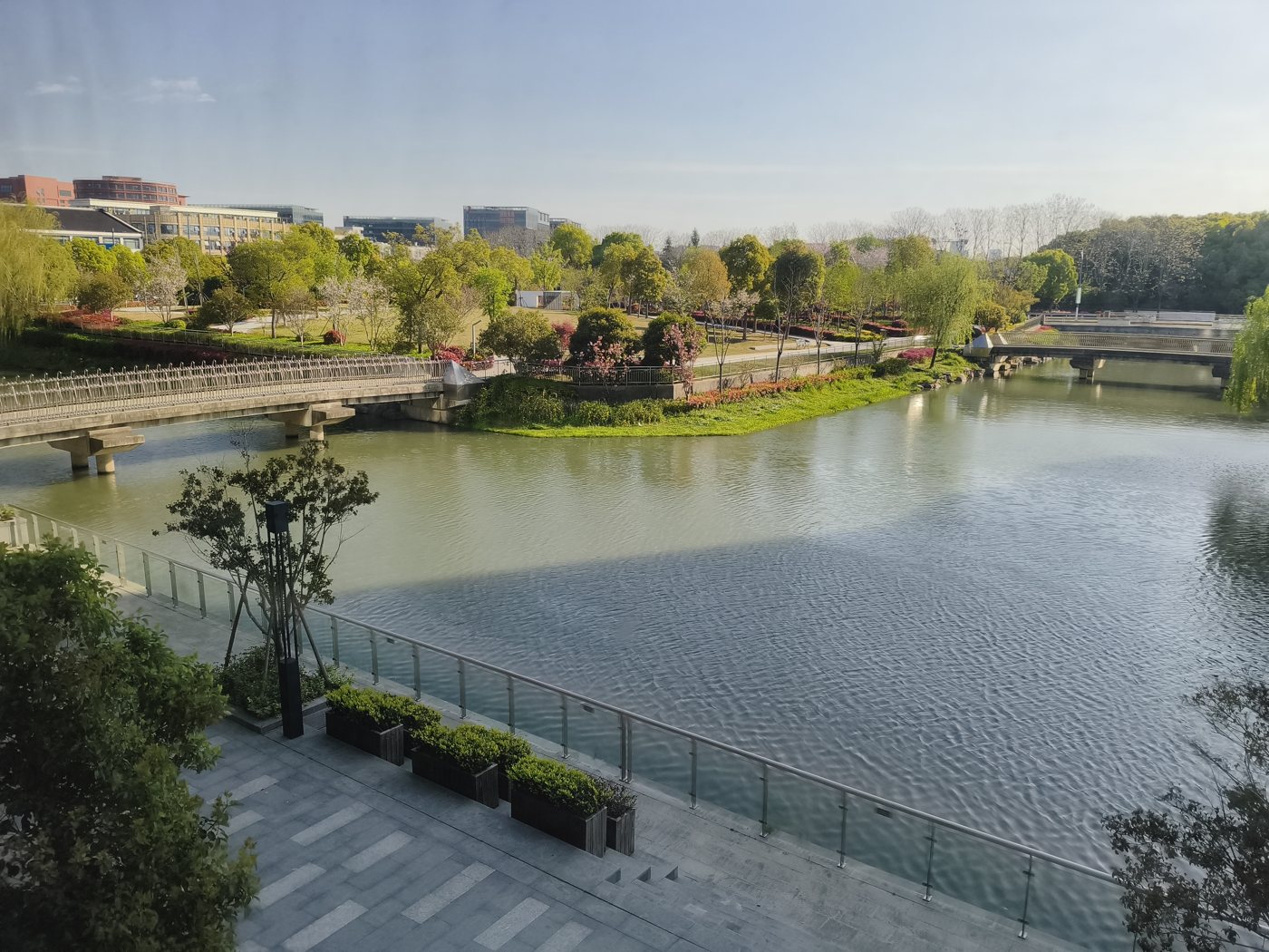 A calm river with a stone bridge, trees and a paved terrace seen from the voco Shanghai Hongqiao Hub lounge window
